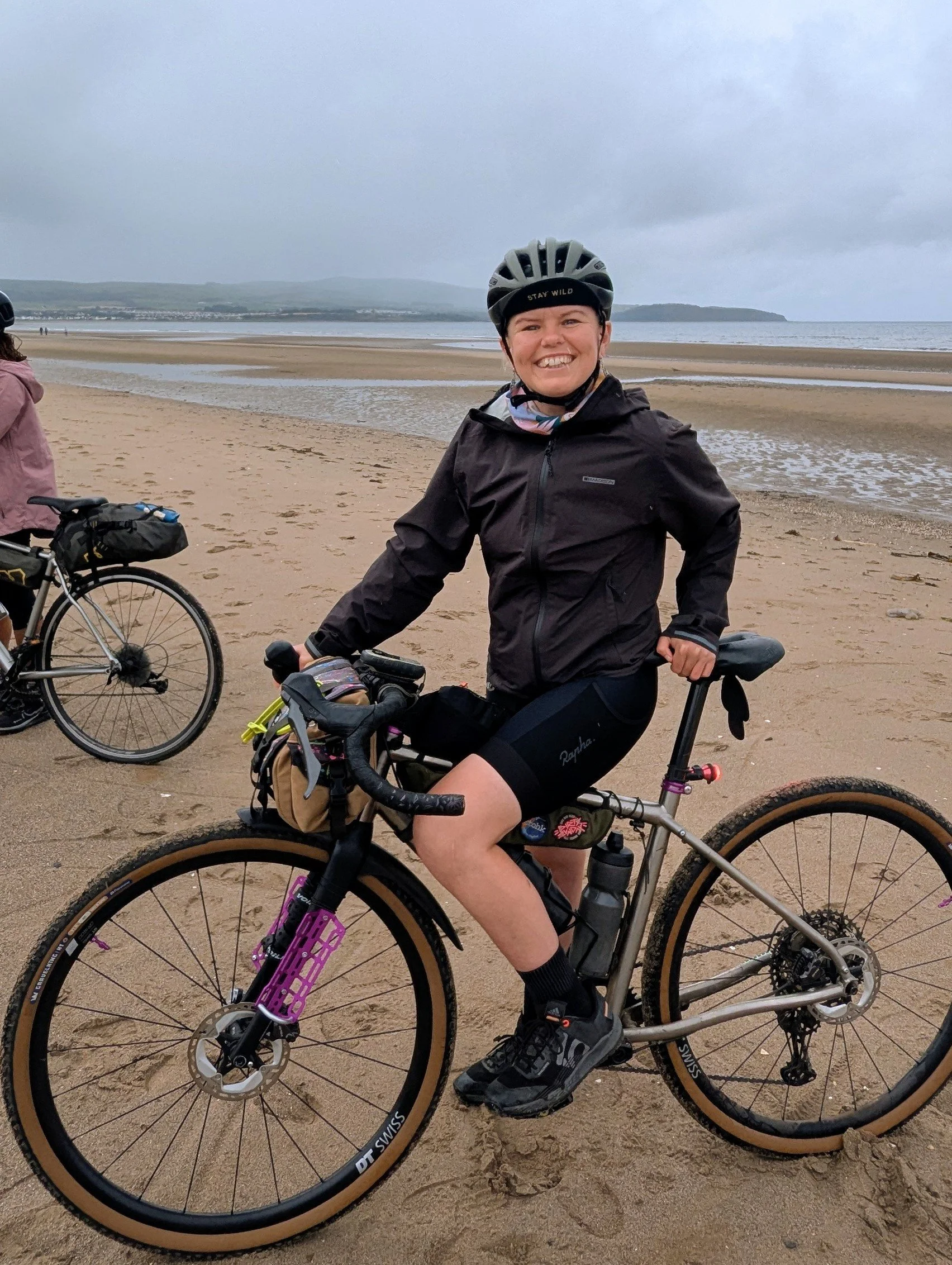 A smiling woman wearing a helmet and cycling gear, standing with her mountain bike on a sandy beach with tide pools, overcast sky, and distant hills in the background.