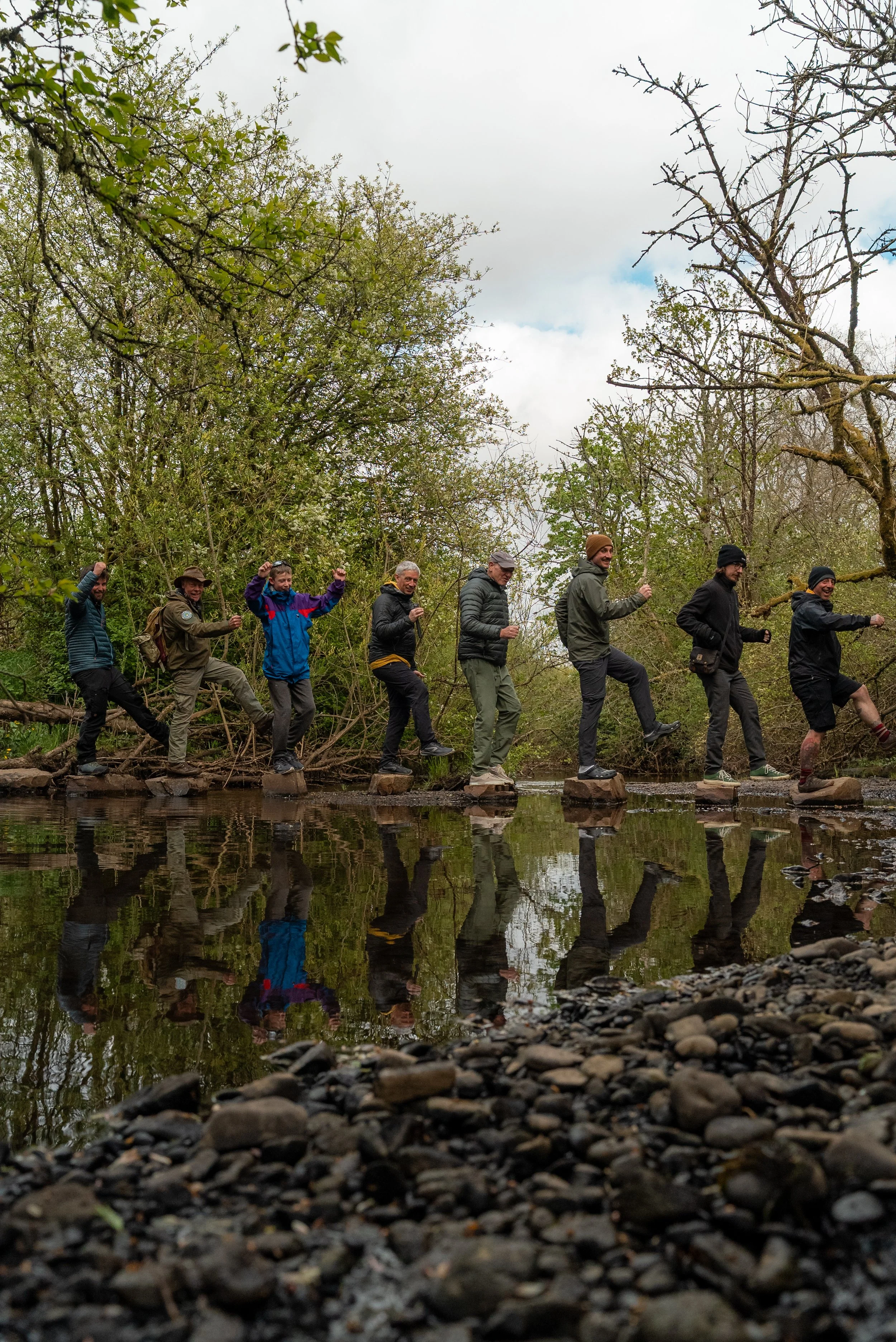 Seven people in outdoor clothing are walking in a line on rocks over a river, surrounded by trees with sparse leaves and cloudy sky.