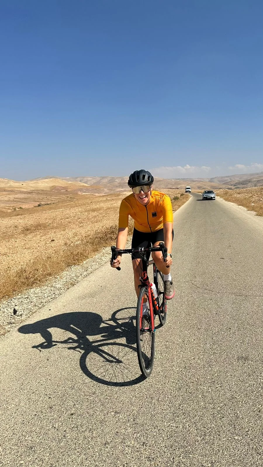 A person cycling on a rural road in a desert landscape under a clear blue sky, wearing a yellow jersey, helmet, and sunglasses.