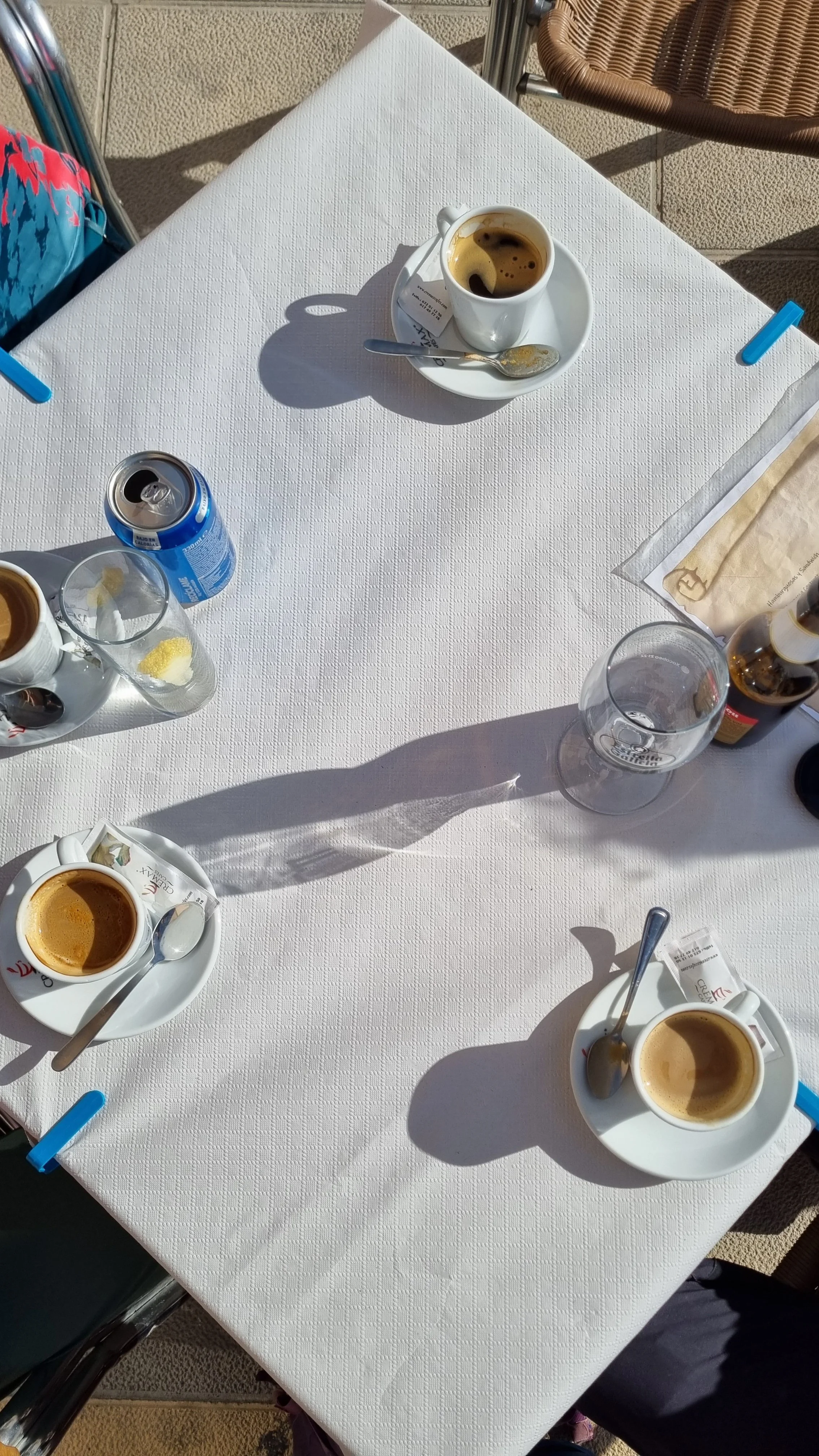 A white outdoor table with three cups of coffee, a can of soda, a glass with lemon slices, and various utensils and papers, with shadows and sunlight on the table.