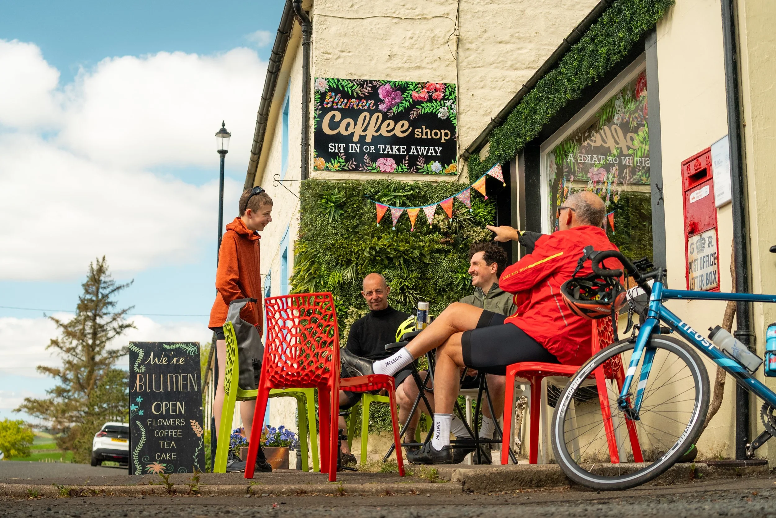 Group of cyclists and friends sitting outside a coffee shop, engaging in conversation, with bicycles parked nearby and colorful chairs. There is a sign indicating the shop offers coffee, tea, cake, and flowers.