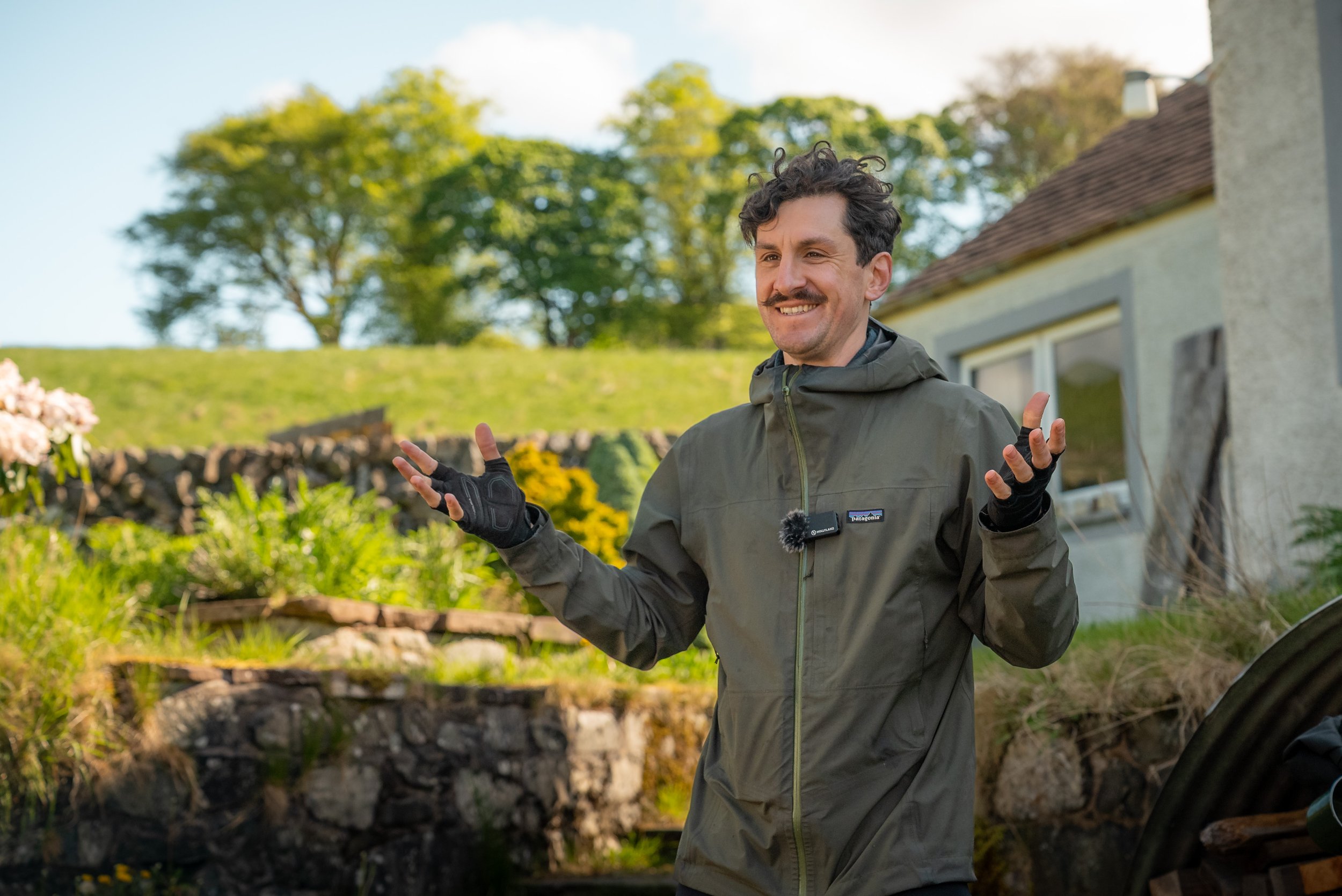 A man with dark curly hair and a mustache smiling outdoors, wearing a gray jacket and black gloves, standing in front of greenery and a house with a sloped roof.