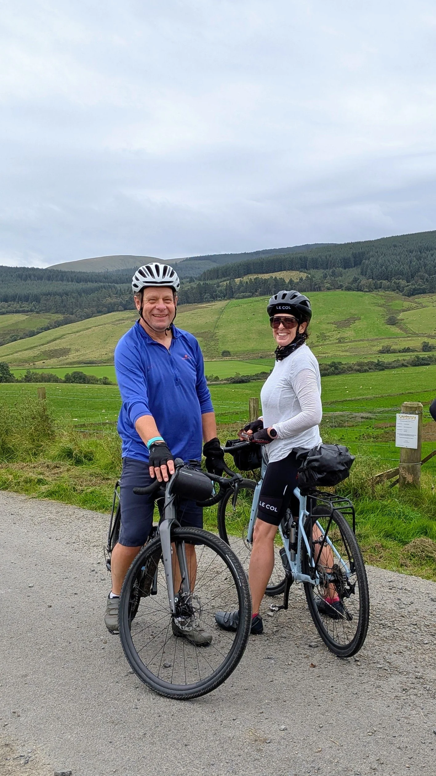 A man and woman wearing cycling helmets and gear, standing with their bicycles in a rural area with green rolling hills and trees in the background.