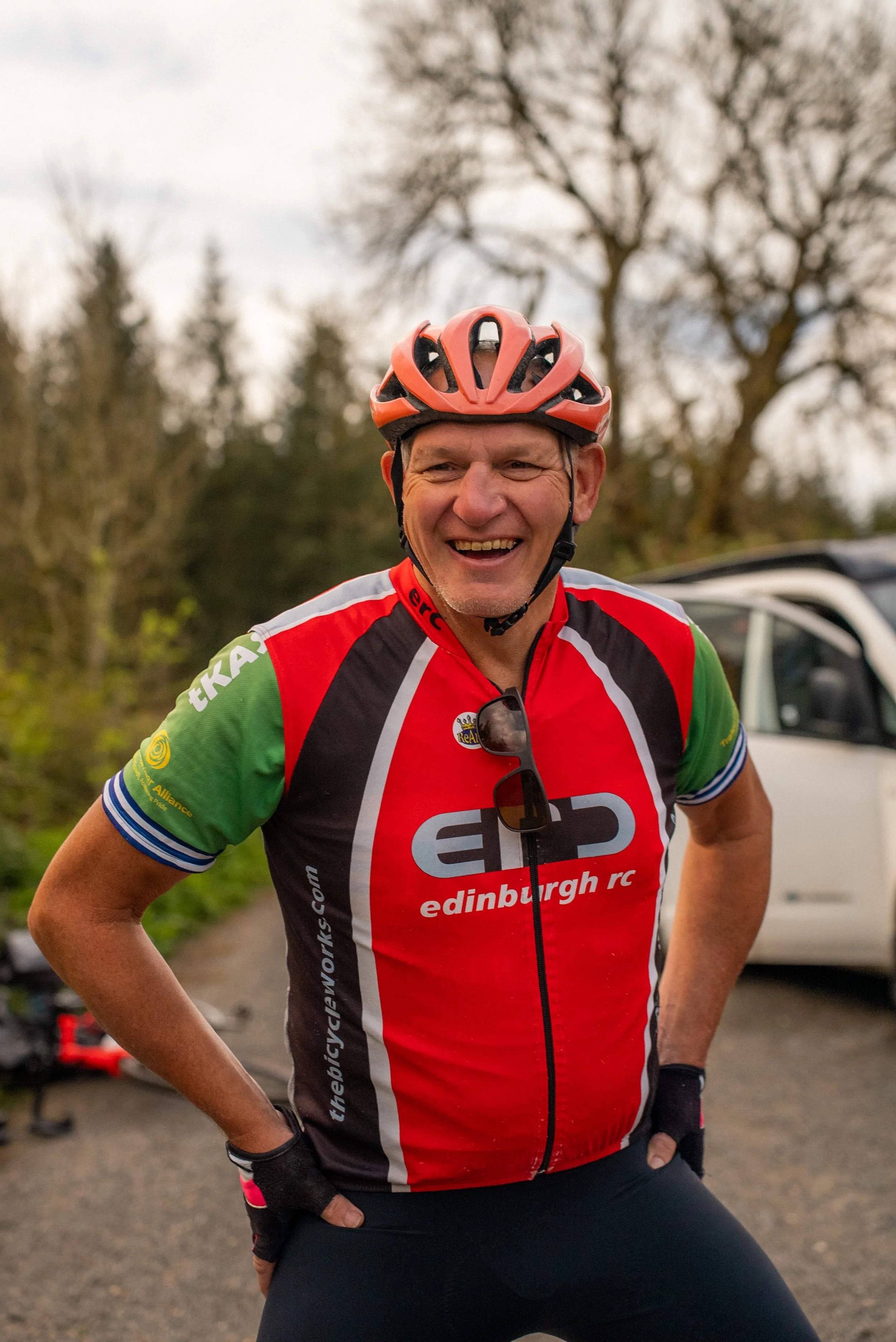 A cheerful man wearing a red helmet and a colorful cycling jersey with the text 'edinburgh rc' smiles outdoors. He stands with hands on hips in front of trees and a vehicle on a cloudy day.
