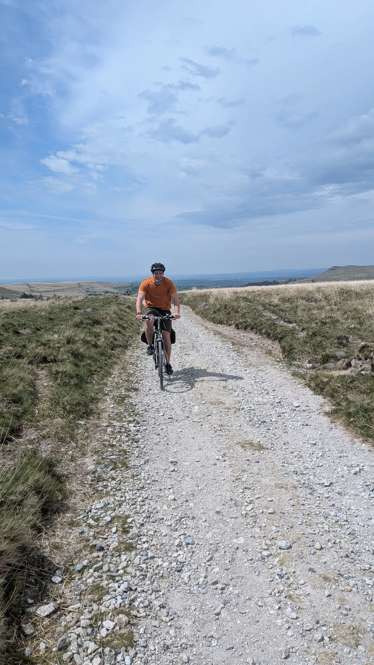 A man biking on a gravel trail in a rural area, wearing a helmet and an orange shirt, with open fields and cloudy sky in the background.
