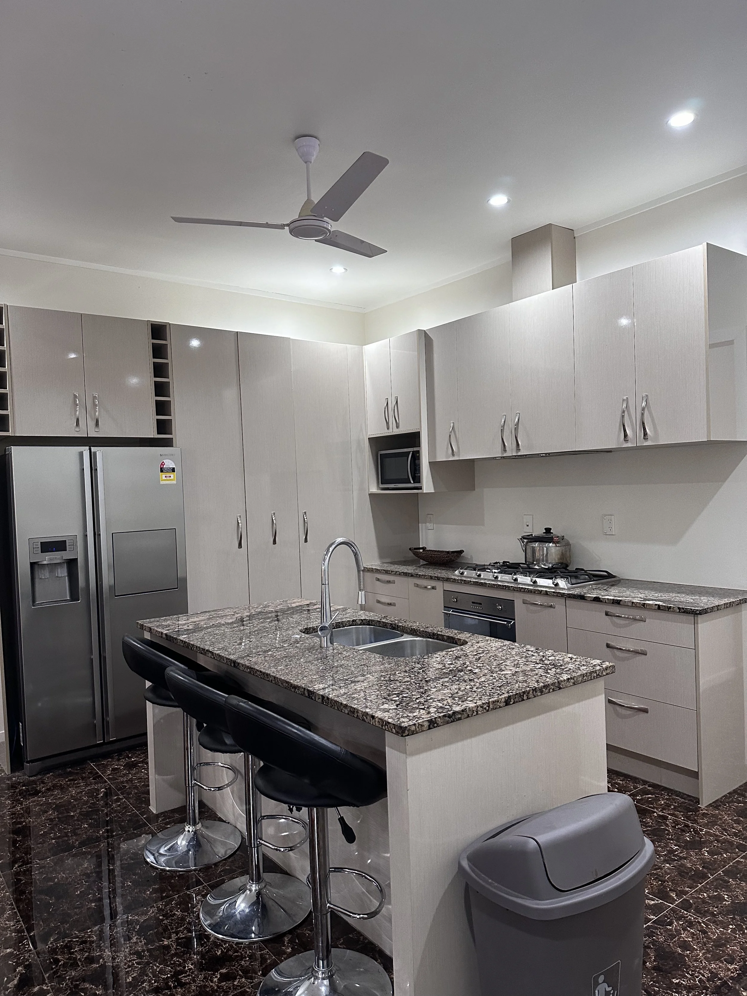Kitchen with beige cabinets, granite countertops, stainless steel appliances, a refrigerator, microwave, gas stove with pot, and a kitchen island with seating. Ceiling fan and recessed lighting are visible.