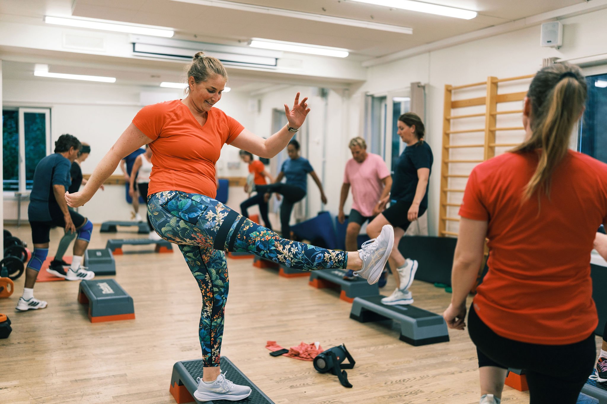 En gruppe mennesker trener i et treningsstudio, der en kvinne i oransje t-skjorte og blomstrete leggings hopper opp på en stepkasse mens andre følger med.