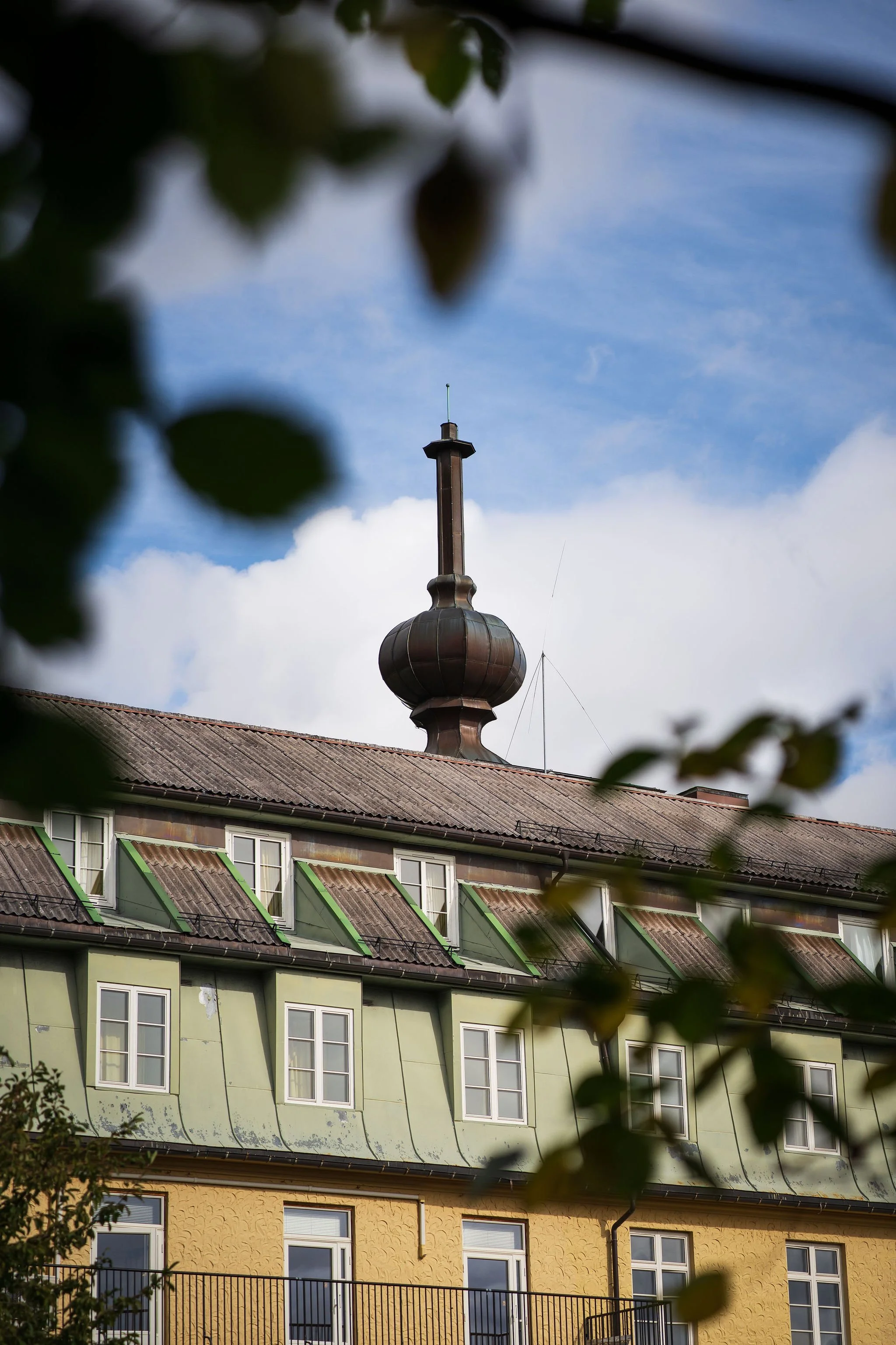 Bilde av en bygning med grønne og gule fasader, tak med takvinduer, og en topp med en cupola og tårn med en antenne. Noen blader er synlige i forgrunnen.