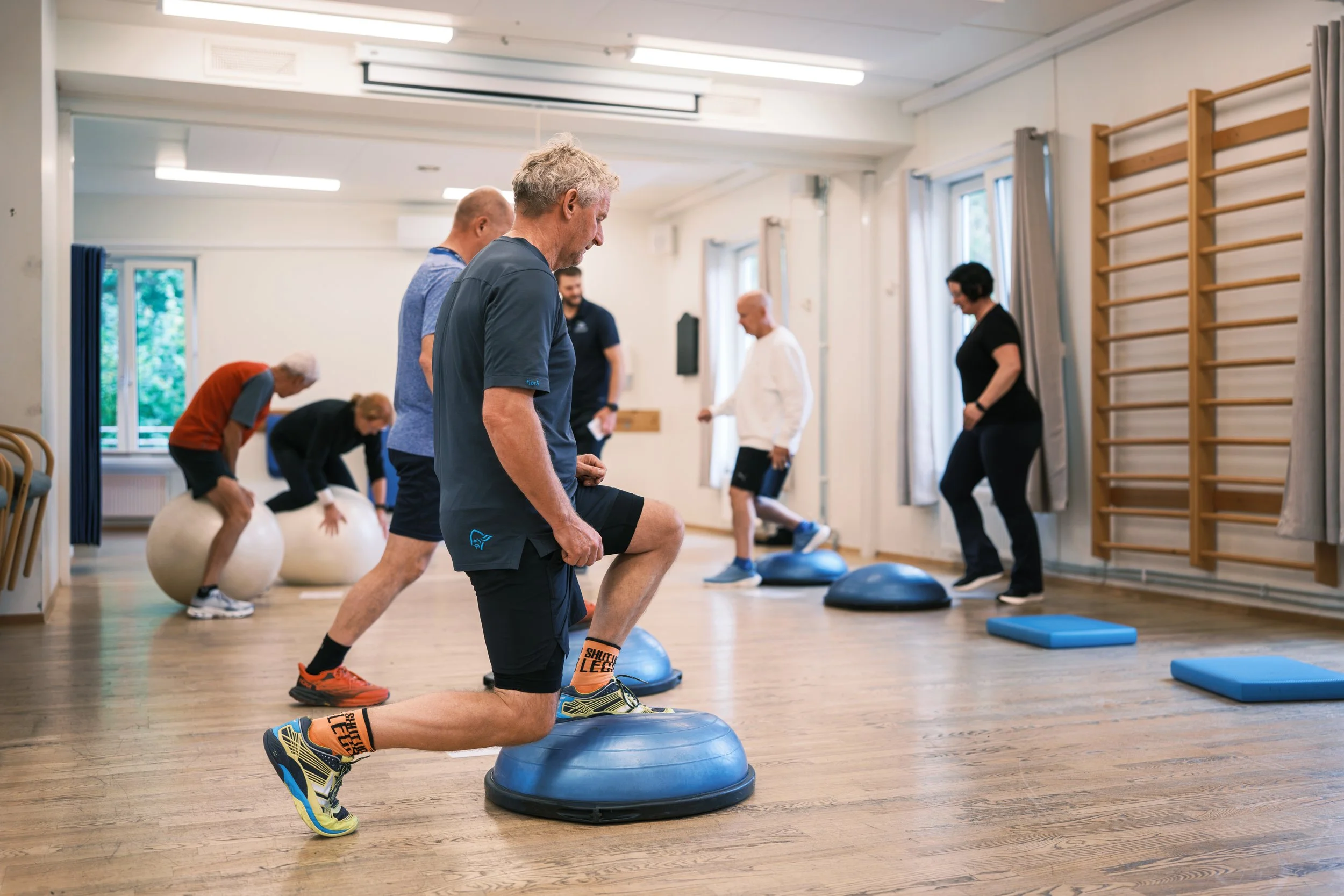 Mann som deltar i en fysisk treningsklasse, knebøyøvelse med balanceball i et treningsrom med andre mennesker som trener.