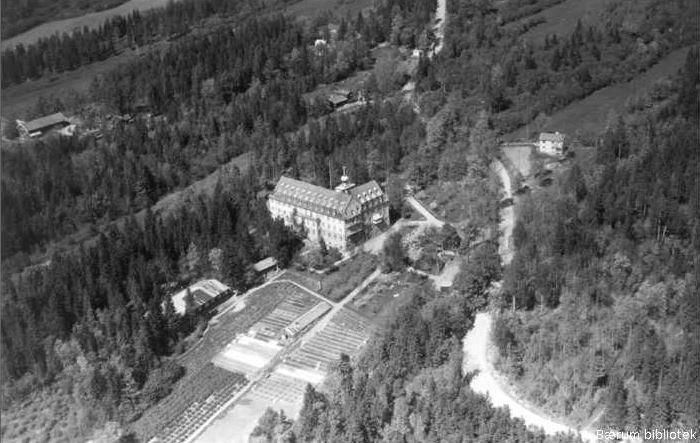 Aerial view of a large building surrounded by dense forest, with greenhouses and a river nearby.