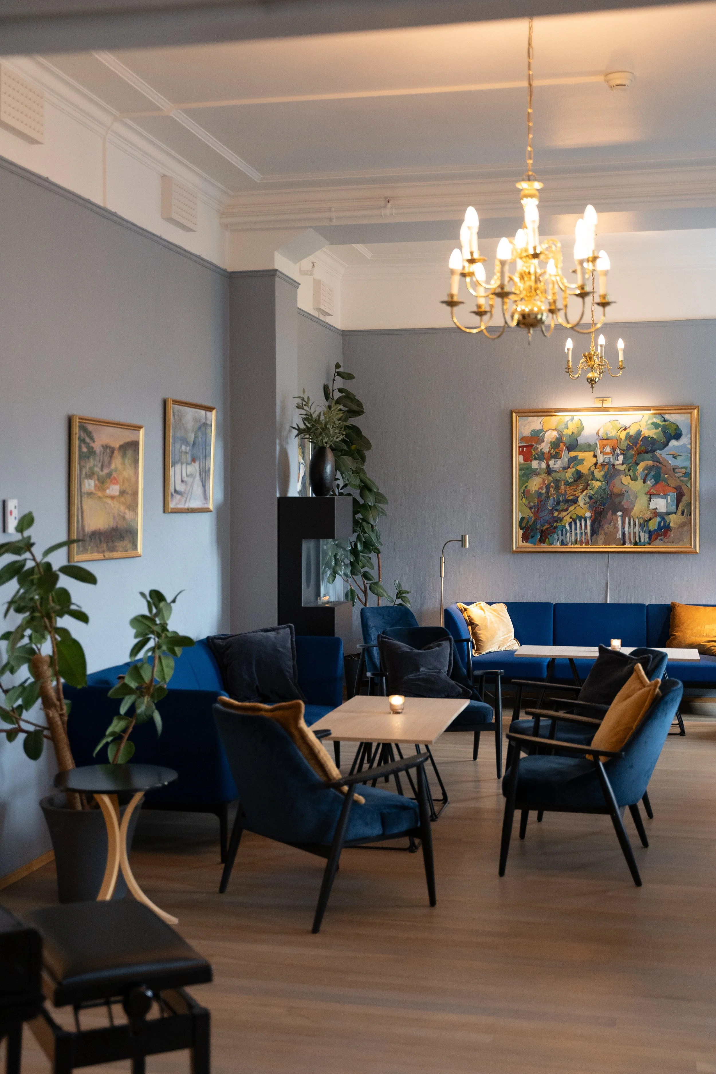 Et rom med blå sofaer og flere stoler rundt små bord, veggdekorasjoner og kunst på veggen, planter og lysarmaturer i taket.