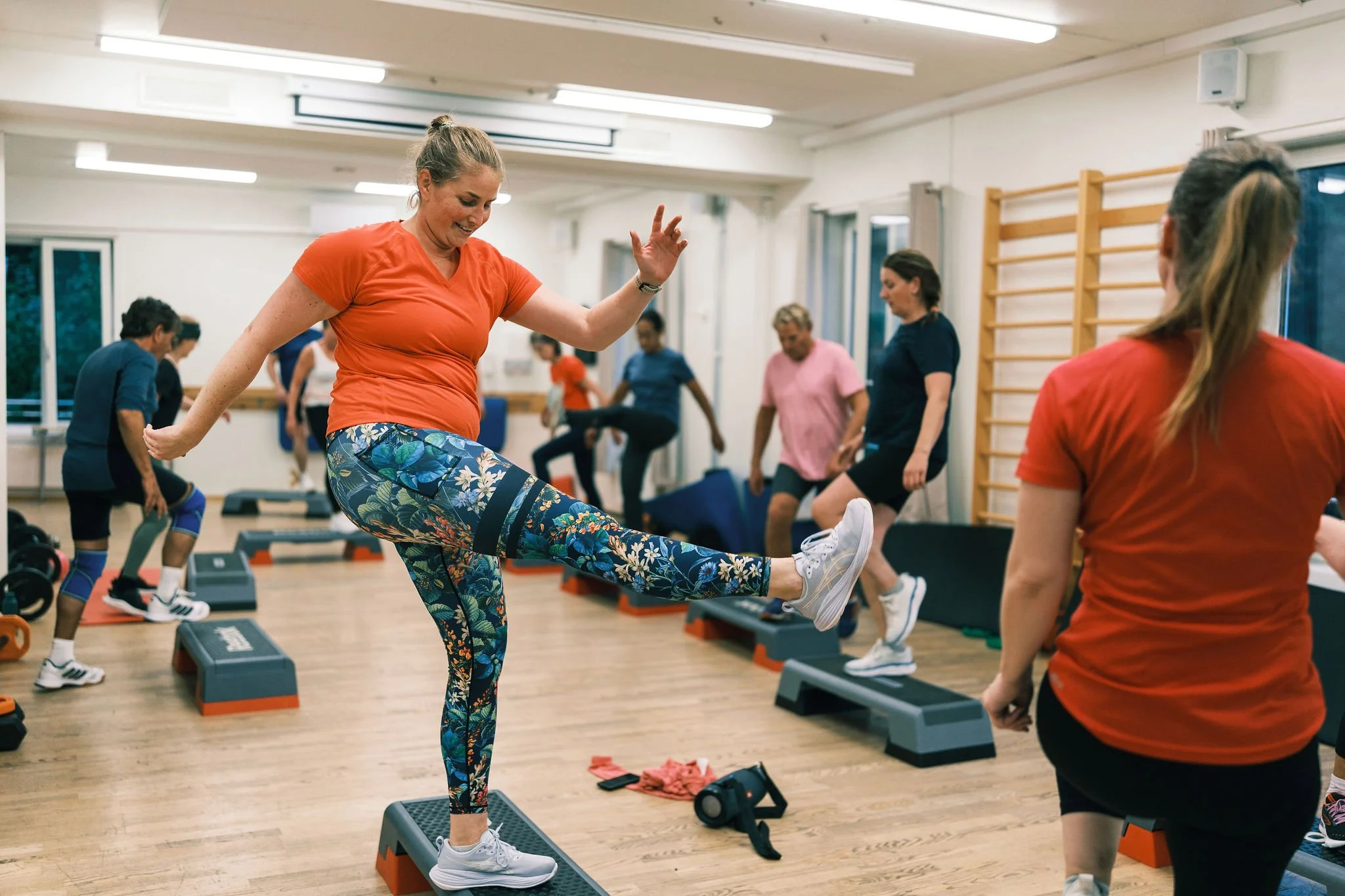 En kvinne i en rosa t-skjorte og fargerike leggings trener balanse på en treningsplate i et treningsstudio, mens andre mennesker trener i bakgrunnen.