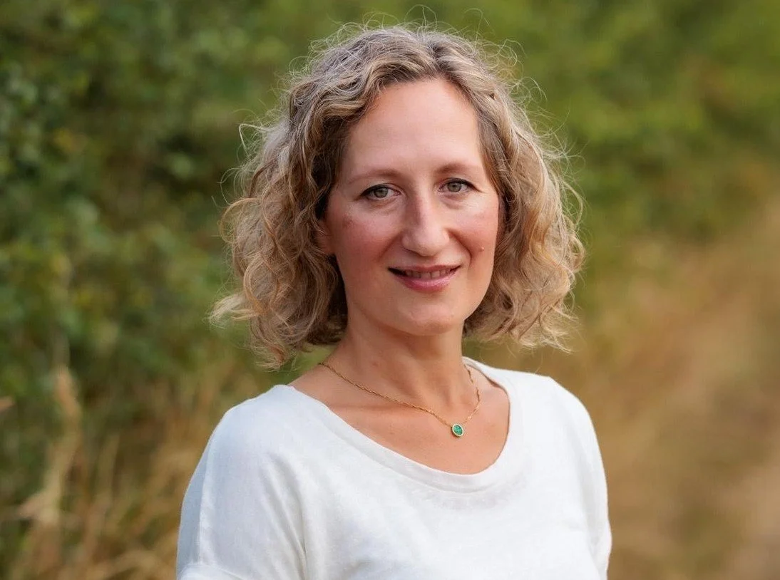 A woman with curly blond hair wearing a white shirt and a gold necklace with a green pendant, standing outdoors with a blurred green and brown background.
