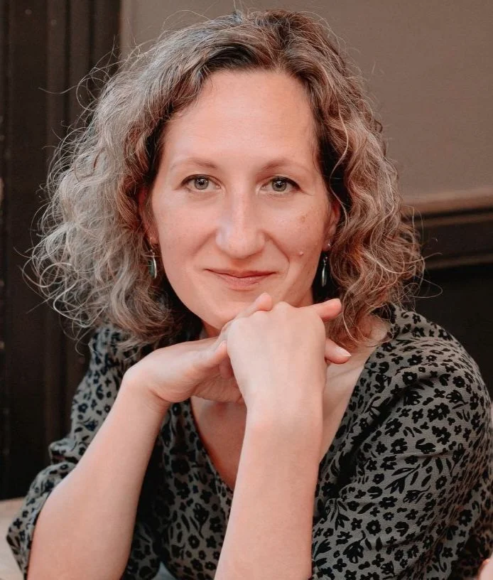 A woman with curly, shoulder-length gray hair, wearing a black and gray patterned top, sitting indoors with her chin resting on her hand, smiling softly at the camera.