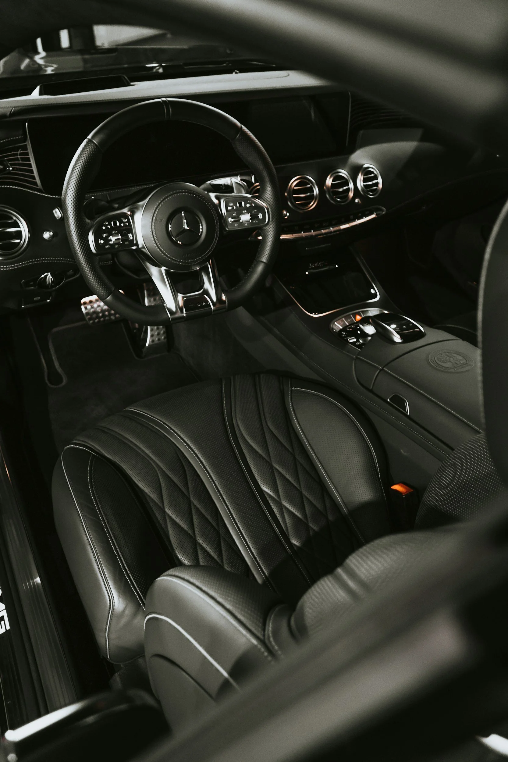 Interior view of a luxury car with a black steering wheel, digital dashboard, three round air vents, and black leather seats with white stitching.