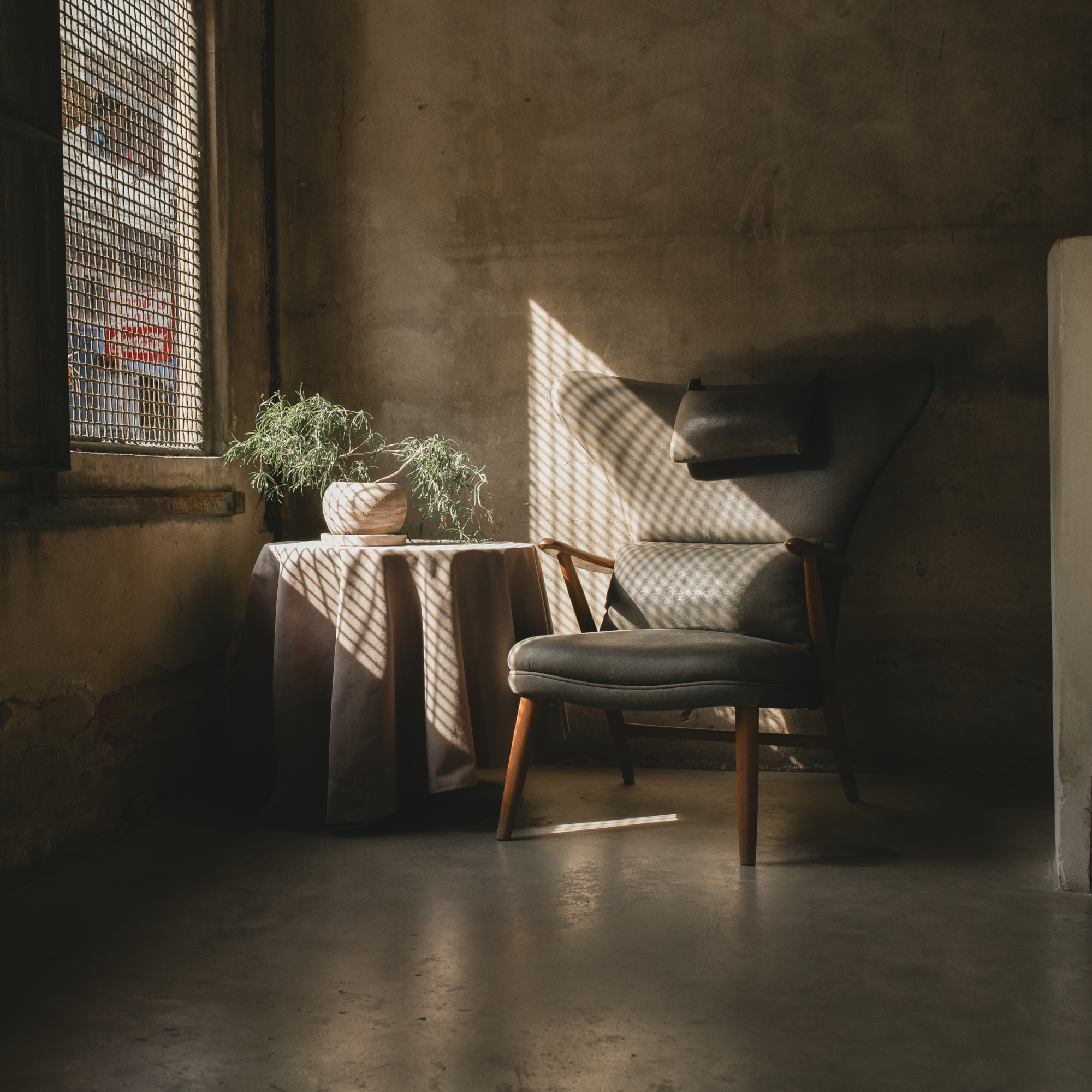 A room with a concrete floor, a wall with cracks, and a window with a metal grid. Sunlight creates shadow patterns on a table and an armchair. There is a potted plant on the table.