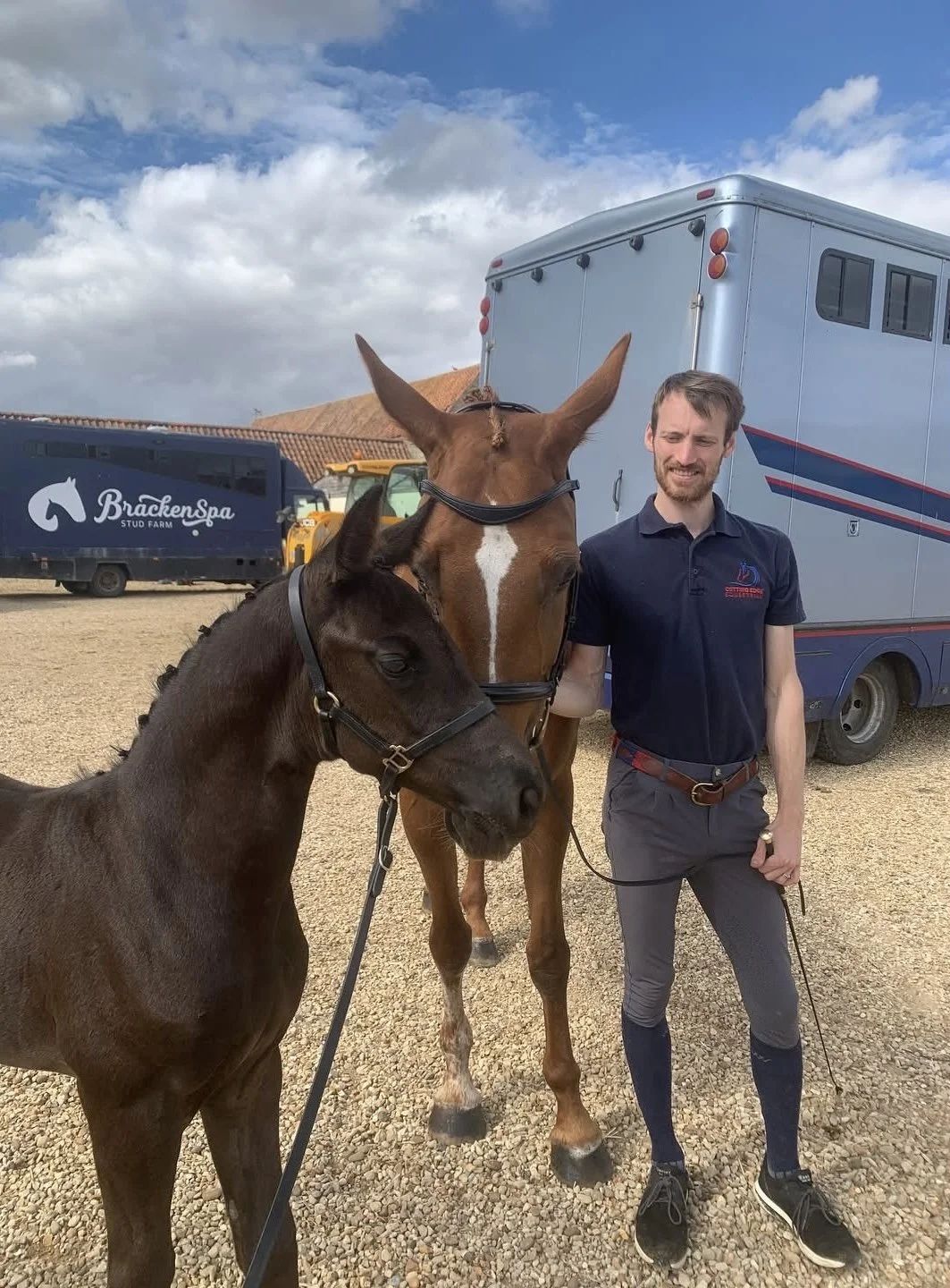 A man standing outside on a gravel surface holding the lead of two horses, one brown and one dark brown, with large horse trailers and a cloudy sky in the background.