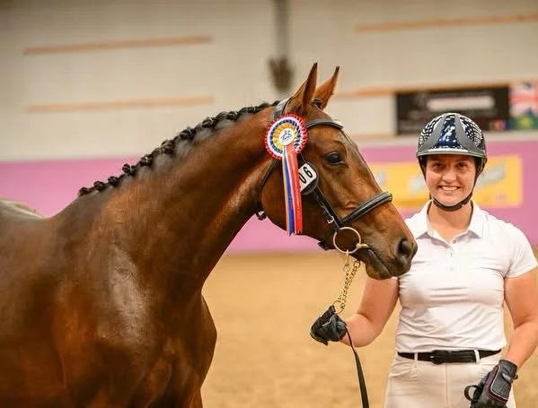 A woman in riding gear standing next to a brown horse with a ribbon on its bridle, inside an indoor riding arena.