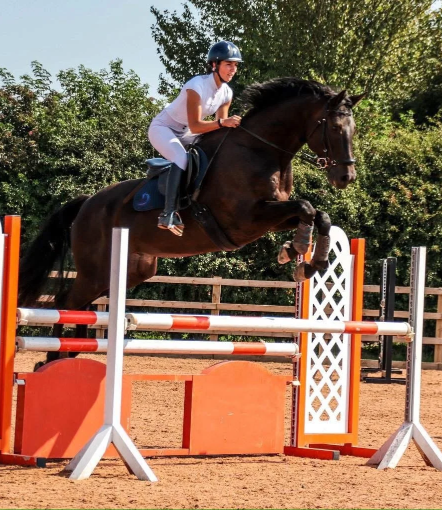 A young woman wearing a helmet and white riding outfit jumping a horse over a hurdle in an outdoor equestrian arena, with trees and a wooden fence in the background.