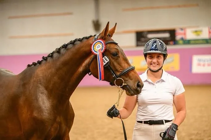 A smiling woman in riding gear holding the bridle of a brown horse with a blue, white, and red ribbon rosette on its bridle, in an indoor equestrian arena.