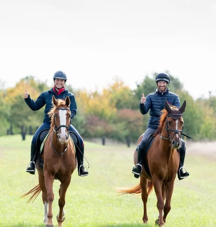 Two people riding horses on a grassy field with trees in the background, giving thumbs up and smiling.