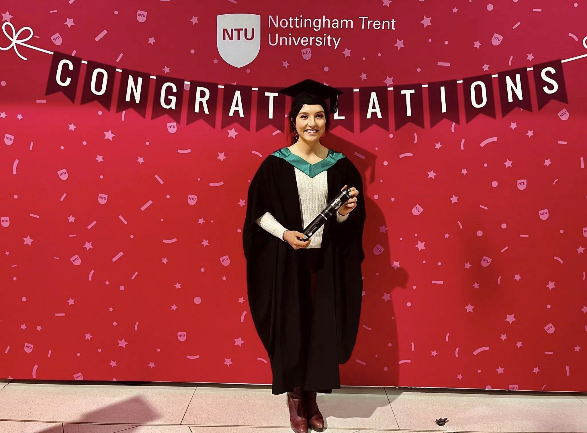 A woman in graduation cap and gown smiling and holding a diploma standing in front of a red backdrop with "Congratulations" banner and Nottingham Trent University logo.
