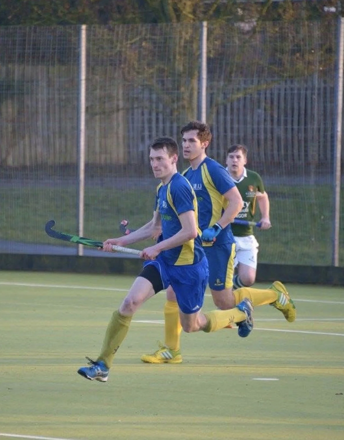 Three male field hockey players running on the field during a game, with one player in a green jersey and two in blue and yellow jerseys, holding field hockey sticks, and a chain-link fence in the background.