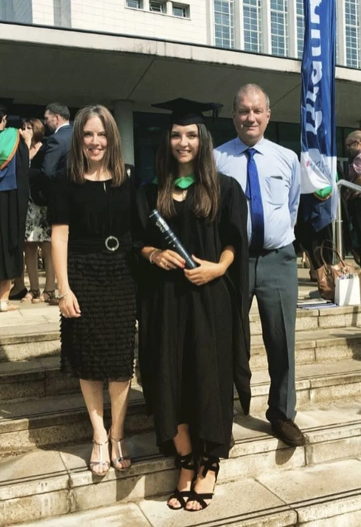 A young woman in a graduation cap and gown holding a diploma, standing on steps with two adults, surrounded by other people in graduation attire outside a building.