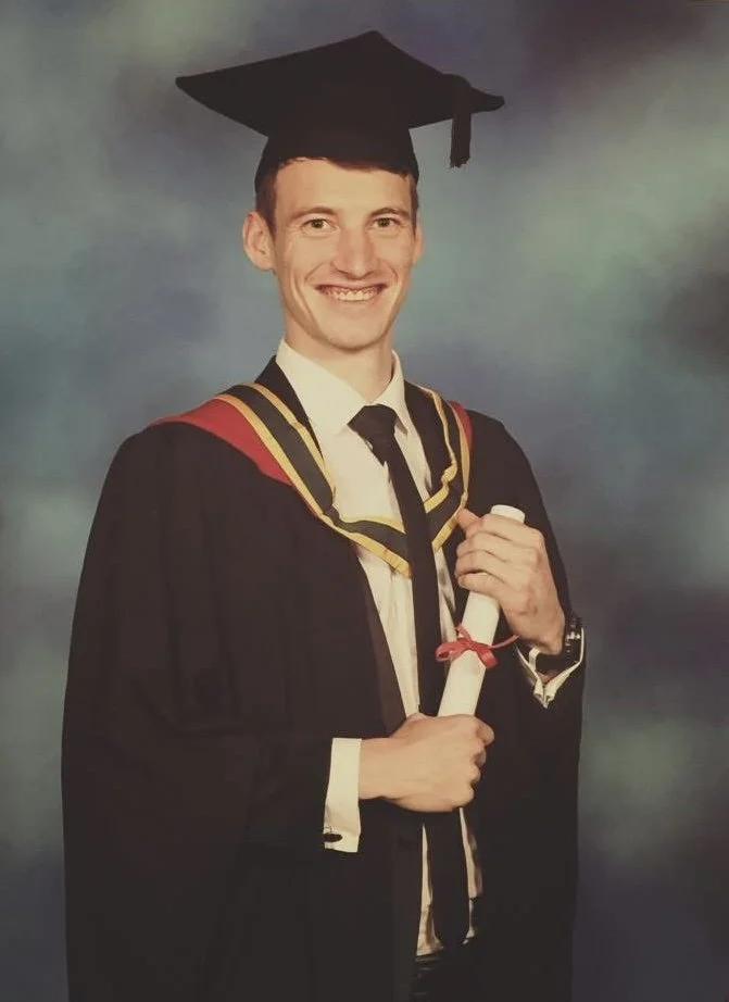 A young man in a graduation cap and gown holding a diploma and smiling at the camera, standing against a blue and green background.
