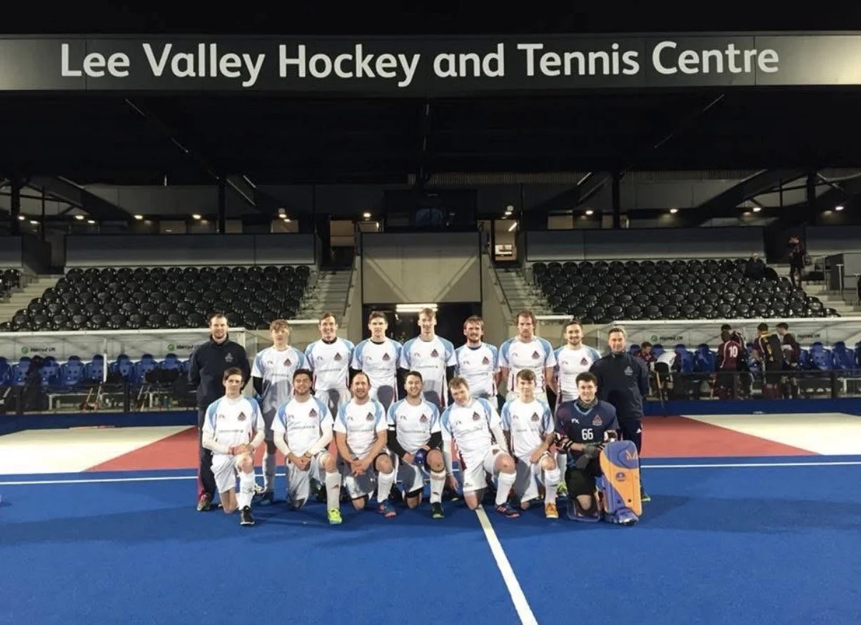 A group of male floorball or hockey players and coaches posing for a team photo on an indoor court at Lee Valley Hockey and Tennis Centre. The team is wearing white and blue uniforms, with one player in black goalkeeper gear. There are empty tiered seating and benches in the background.