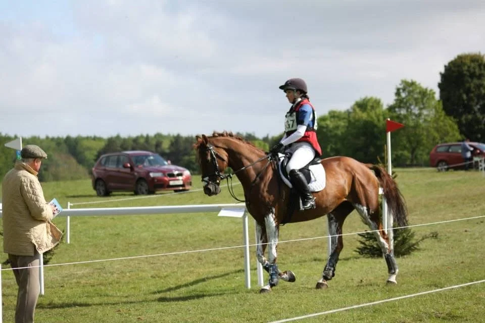 A horse and rider participating in an equestrian event, with a person observing and taking notes nearby on a grassy field, with cars visible in the background.