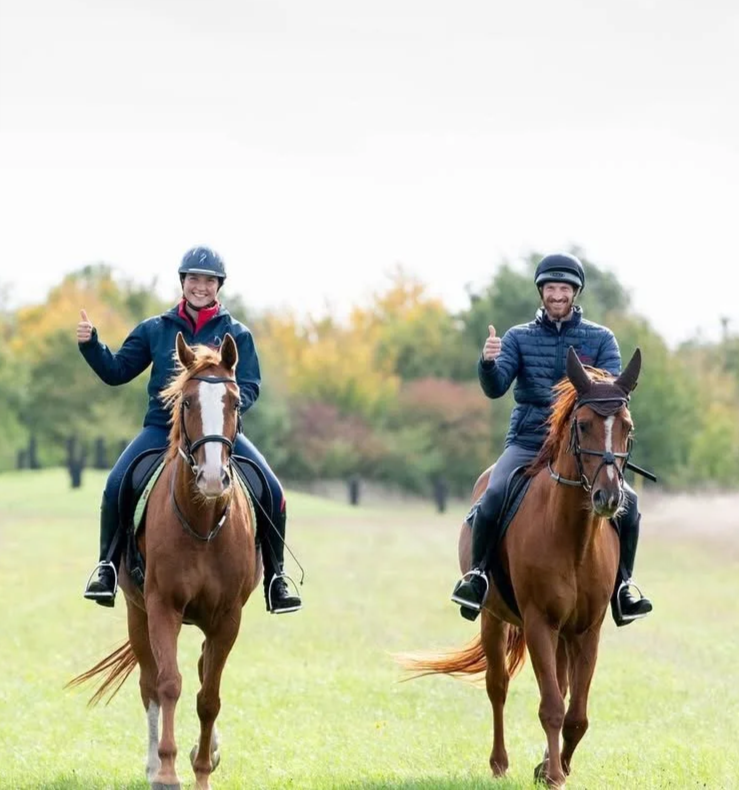 Two people riding horses in a grassy field, giving a thumbs-up and smiling, with trees in the background.