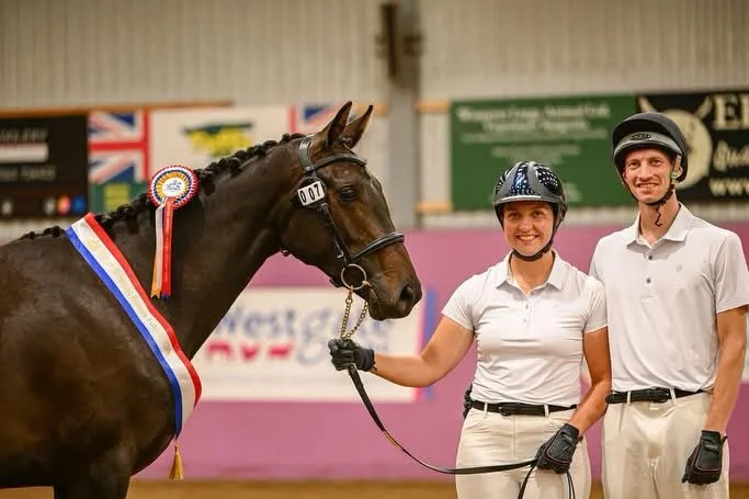 A woman and a man in equestrian attire, standing with a horse decorated with a rosette, inside an indoor riding arena decorated for a competition.