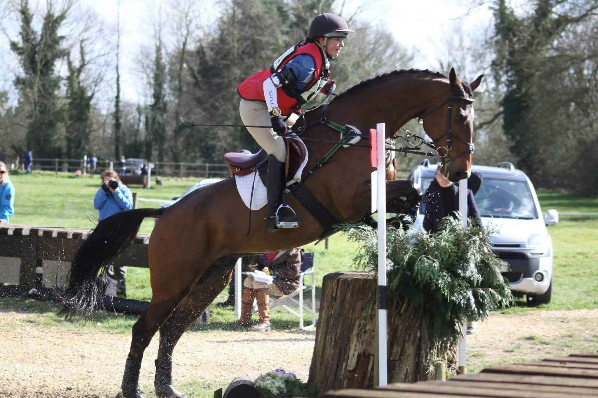 Horse and rider jumping over a log obstacle in an equestrian competition, with spectators and photographers watching in a grassy outdoor arena.