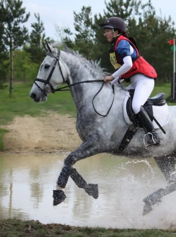 Young female equestrian riding a gray horse through water at a cross-country competition.