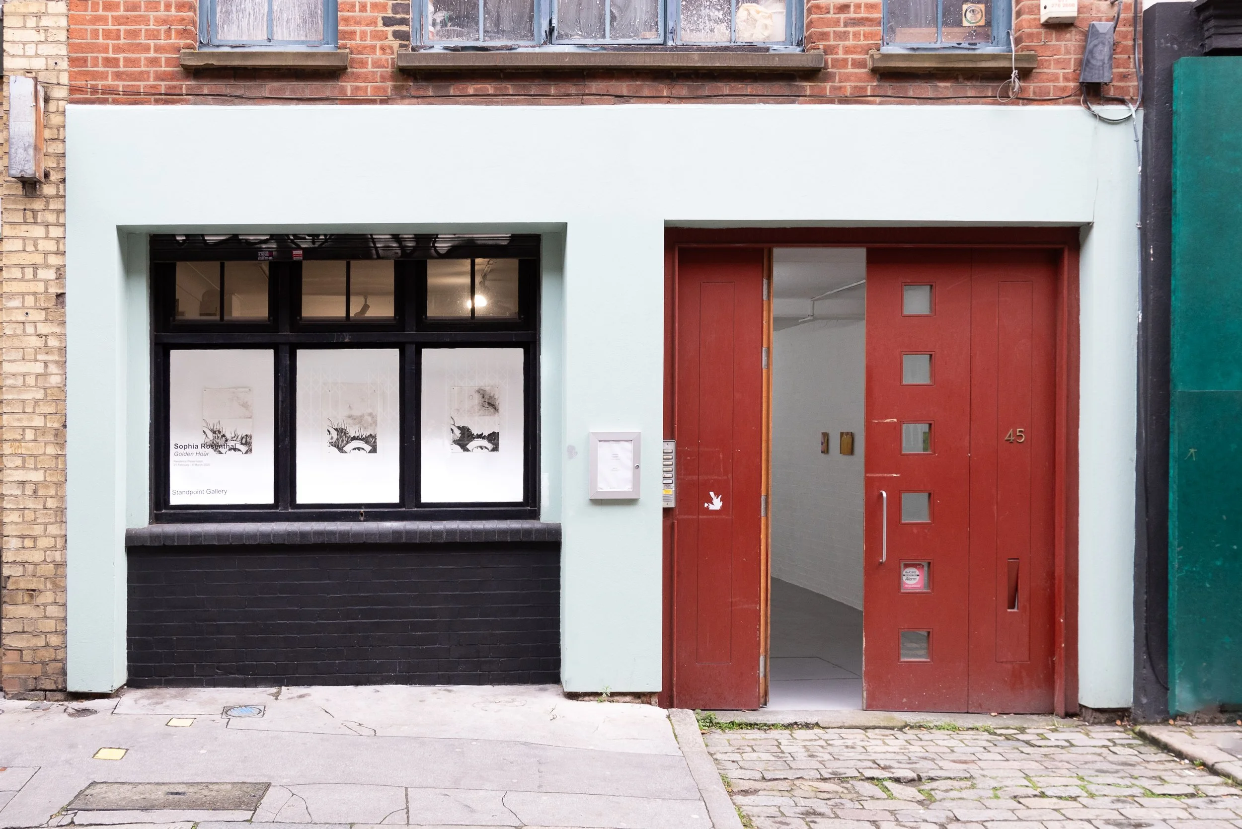 The image shows a storefront with a black-framed window on the left and a red wooden door on the right. The building has exposed brick on the upper part and a painted light mint green on the lower part around the entrance. There is a small sign in the window that reads 'Sophia Rosenthal Golden Hour Standpoint Gallery.' The street in front has cobblestones and sidewalk paving, with some dirt and small patches of plants near the base of the building.