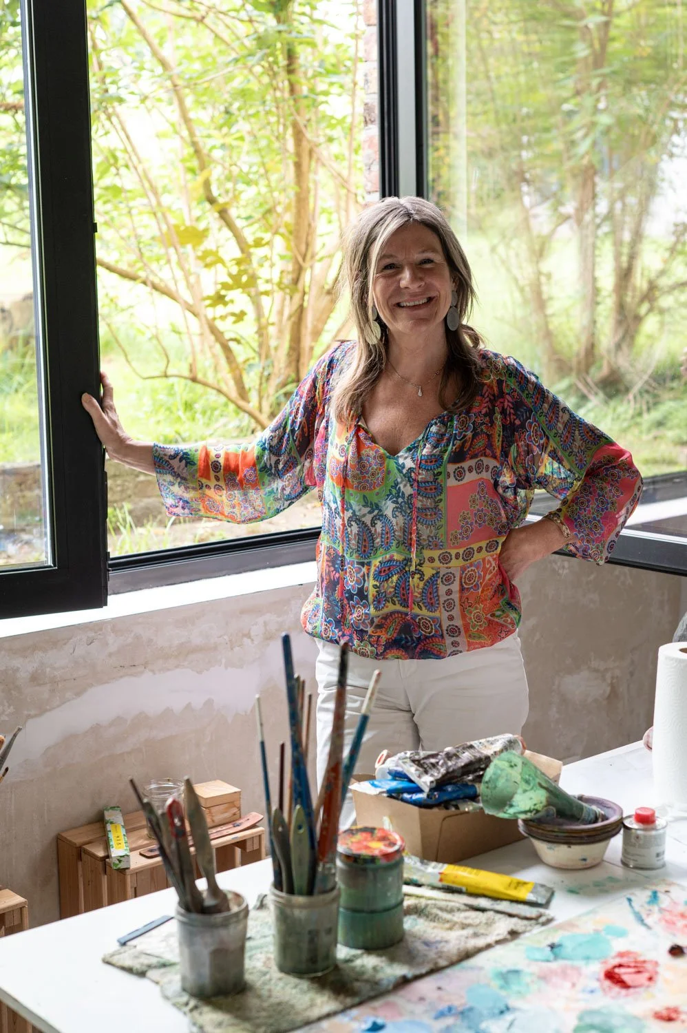 Martine Beyers van Vitavizion die naast een open raam staat met een groene tuin op de achtergrond, in een artistieke ruimte met verfmaterialen op tafel.