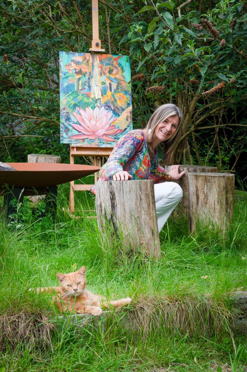 Martine Beyers die op een houten stam zit in een tuin, met een kleurrijk abstract kunstwerk op een ezel achter haar. Een oranje kat ligt in het gras voor haar.
