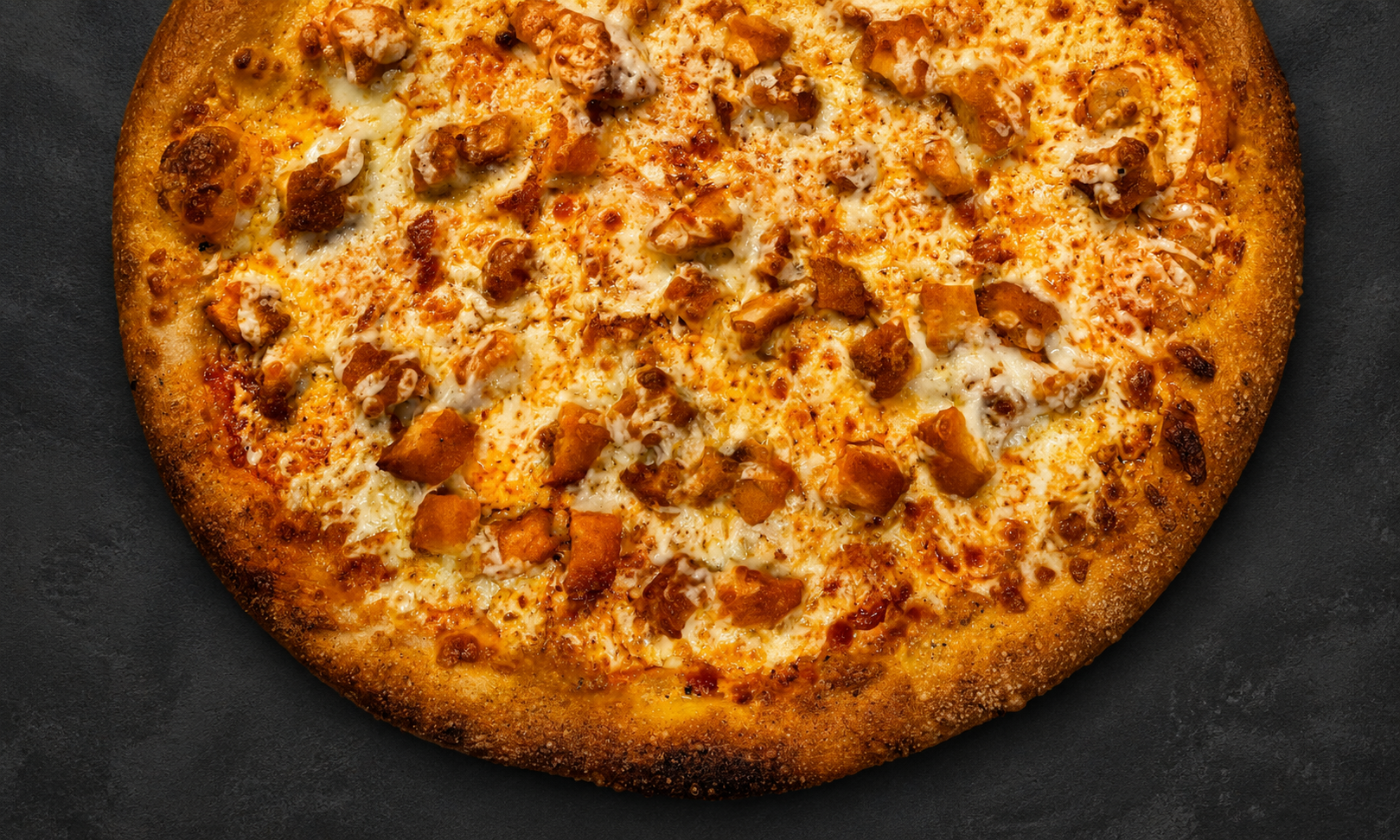 Close-up of a cooked pizza with cheese and chunks of chicken on a dark surface.