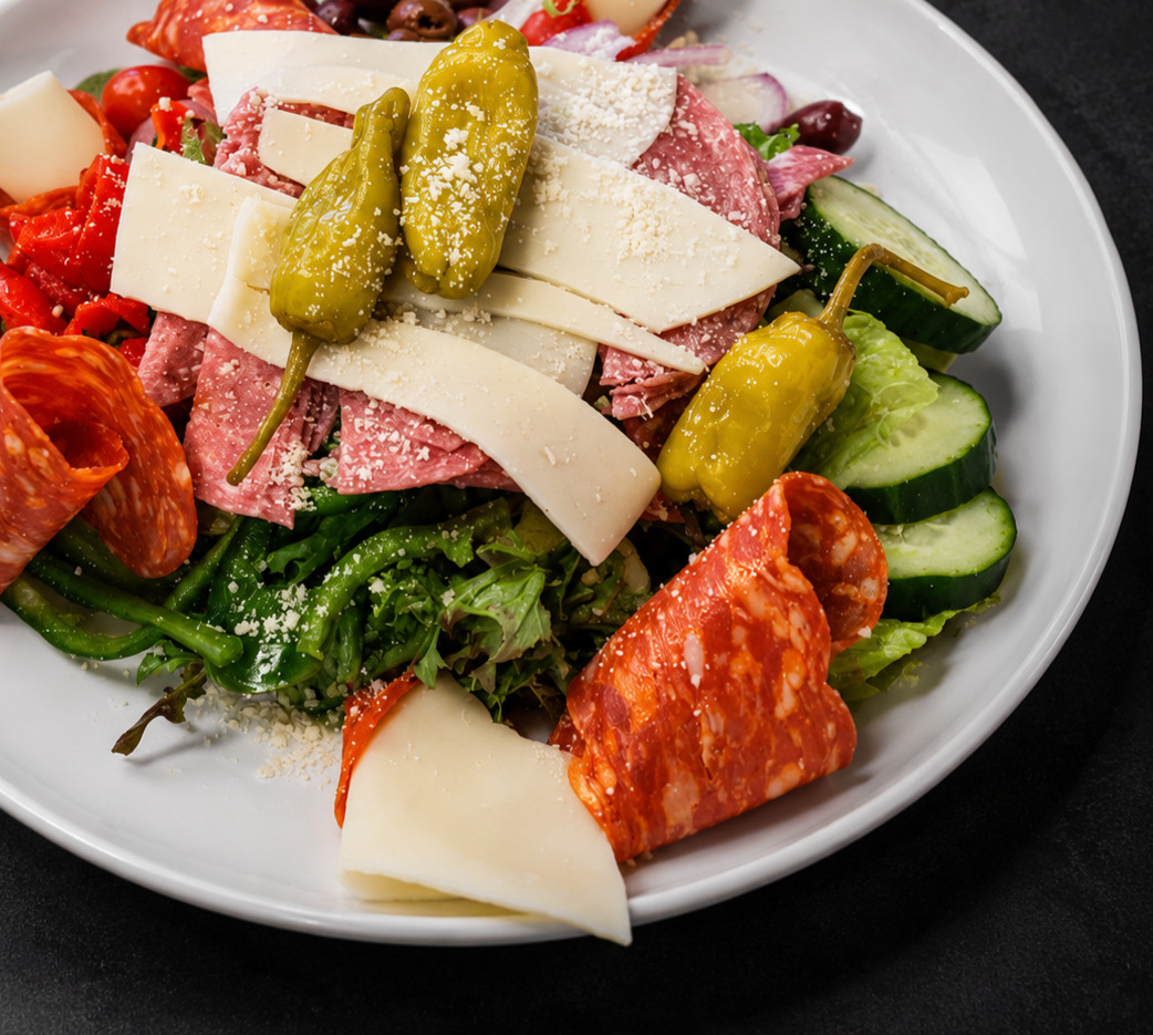 Close-up of an Italian pasta salad containing green pasta, cherry tomatoes, cucumber slices, cheese, pickled peppers, red onions, salami, and shredded cheese in a white bowl.