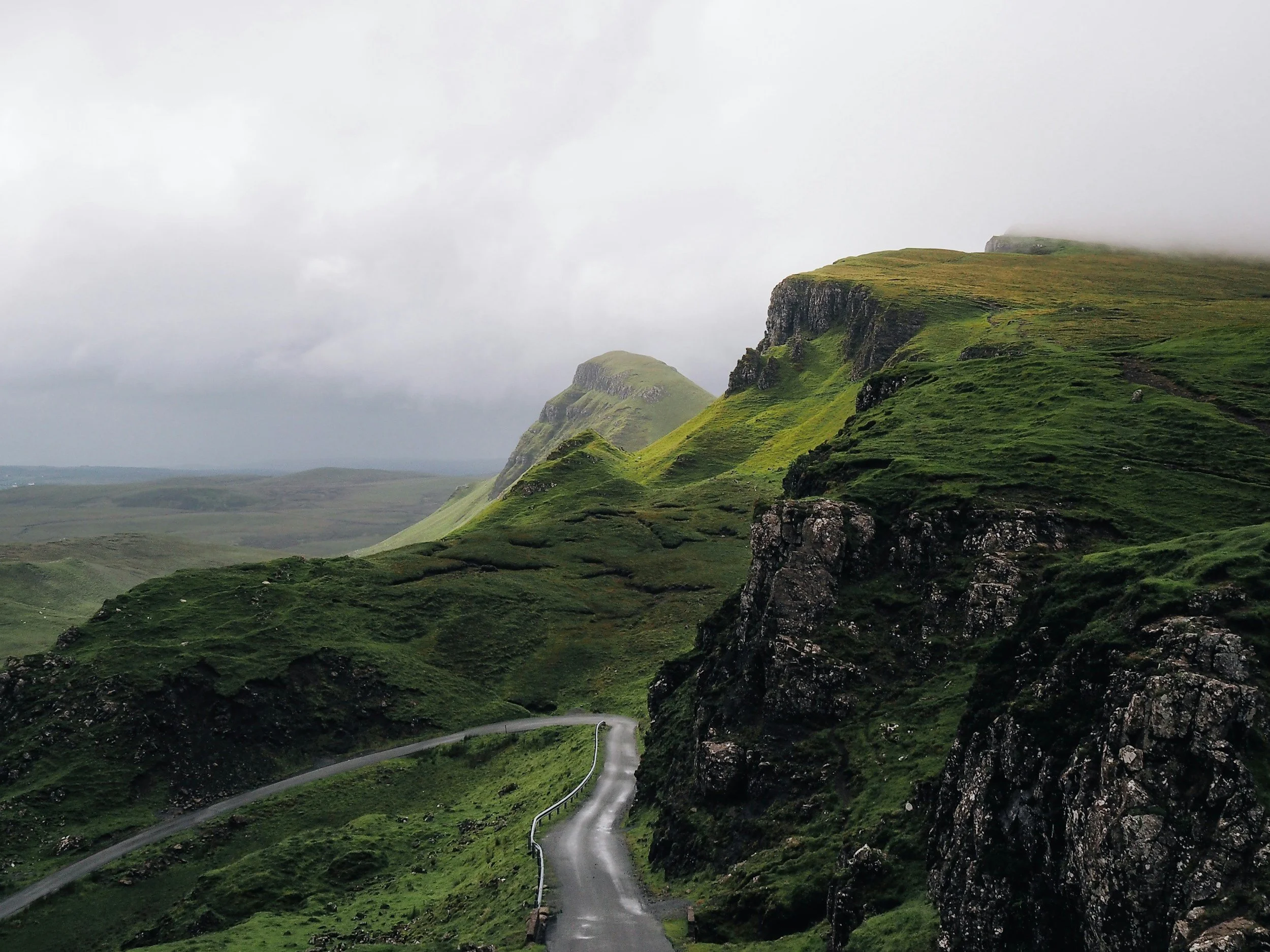 A winding road through green hills and rocky cliffs on a foggy day.