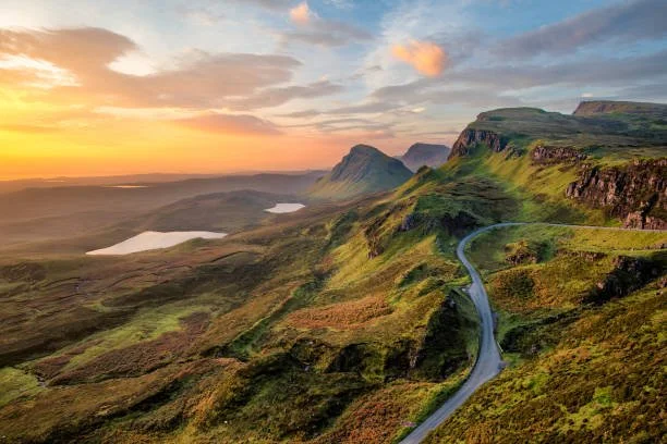 Scenic landscape of rolling green hills with a winding road, distant lakes, and cliffs at sunset.