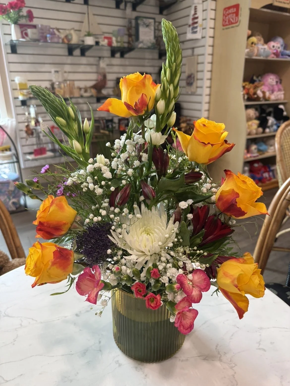 Un bouquet de fleurs colorées dans un vase vert sur une table en marbre, avec des roses jaunes et orange, des œillets roses et blancs, une grande fleur blanche, et des lys blancs en bouton.