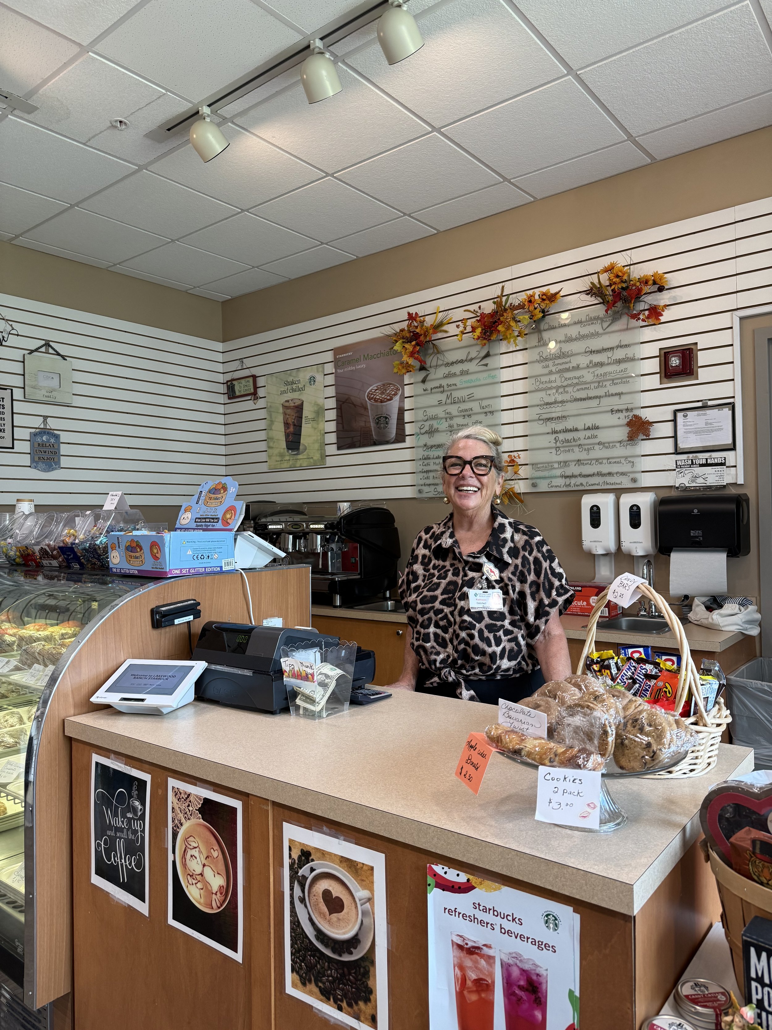 Une caissière souriante derrière le comptoir d'une boutique Starbucks, avec des décorations automnales en haut et un panneau de menu derrière elle, des étiquettes de prix sur les produits en vente, notamment des cookies et des boissons.