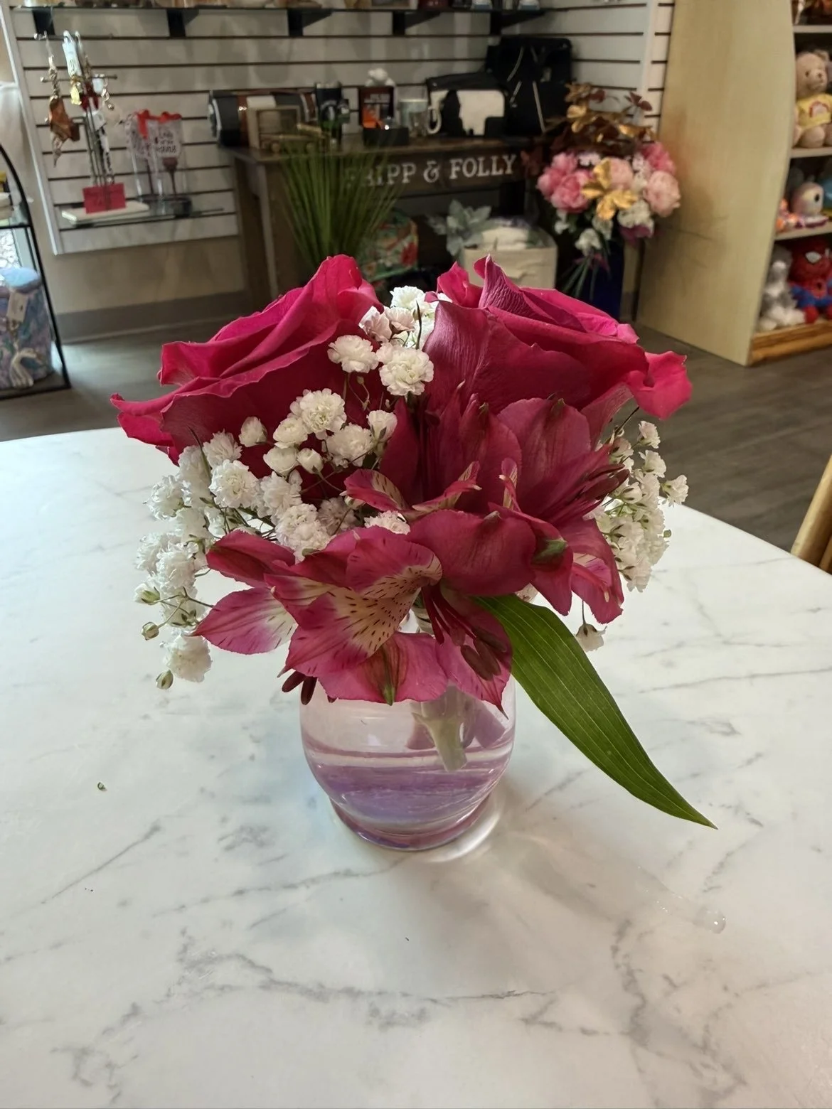 Un arrangement floral avec des roses rouges, des alstroemérias roses et des gypsophiles blanches dans un vase en verre rose pâle sur une table en marbre blanc.