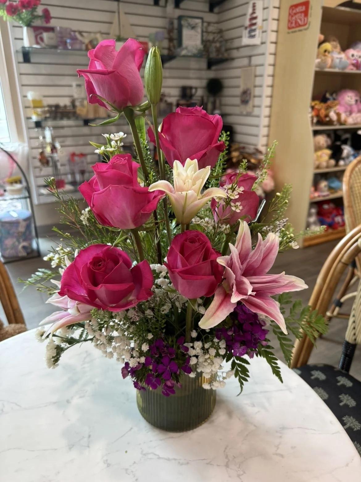 Un arrangement floral avec des roses roses, des lys, et d'autres petites fleurs dans un vase vert, posé sur une table en marbre