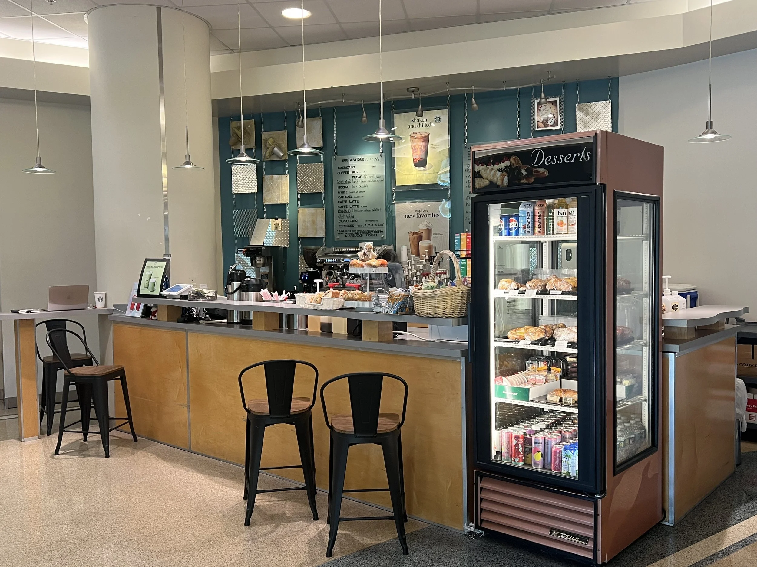 Coin café avec comptoir en bois, chaises en plastique noir, vitrine réfrigérée avec boissons et pâtisseries, décor moderne avec lampes suspendues, tableau de suggestions et affiches de boissons.