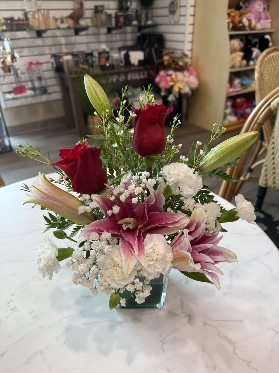 Un arrangement floral avec des roses rouges, des lys roses, des lis blancs et du gypsophile blanc, dans un vase en verre sur une table en marbre.