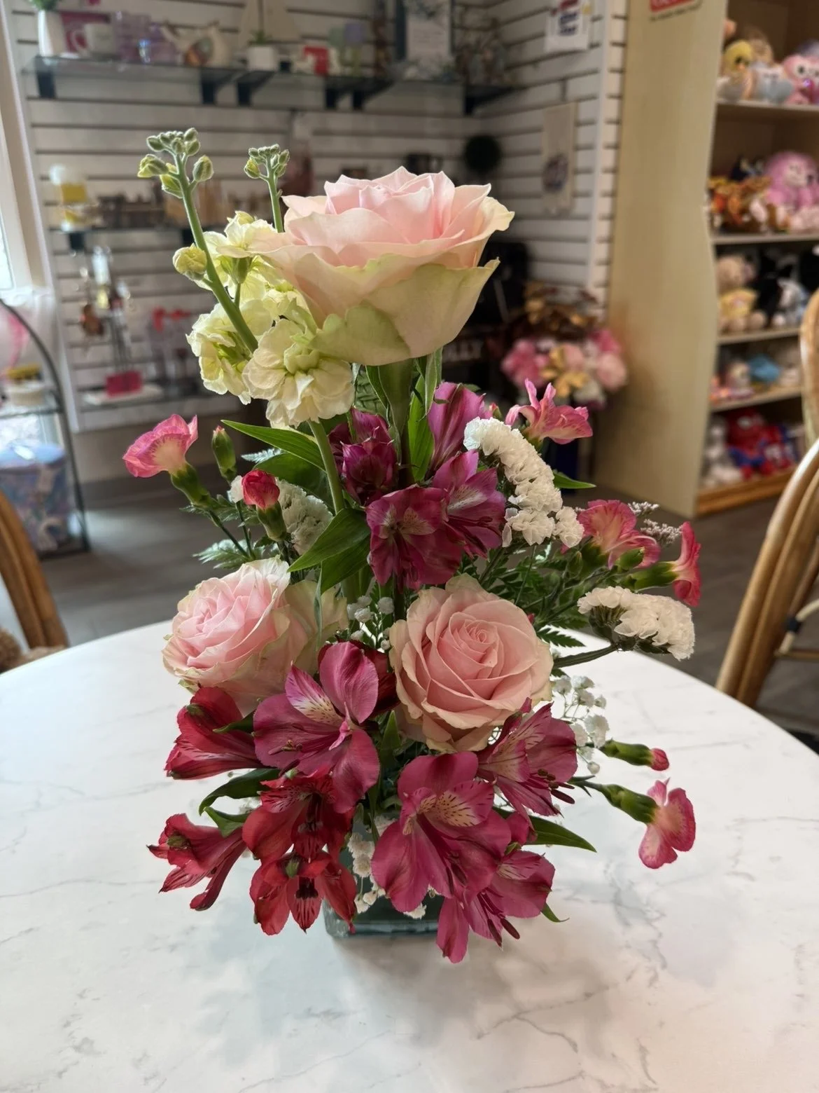 Un vase avec un arrangement floral composé de roses roses, d'alstroemeria roses, de cymbidiums roses, de fleurs blanches et de verdure, placé sur une table en marbre blanc dans une boutique.