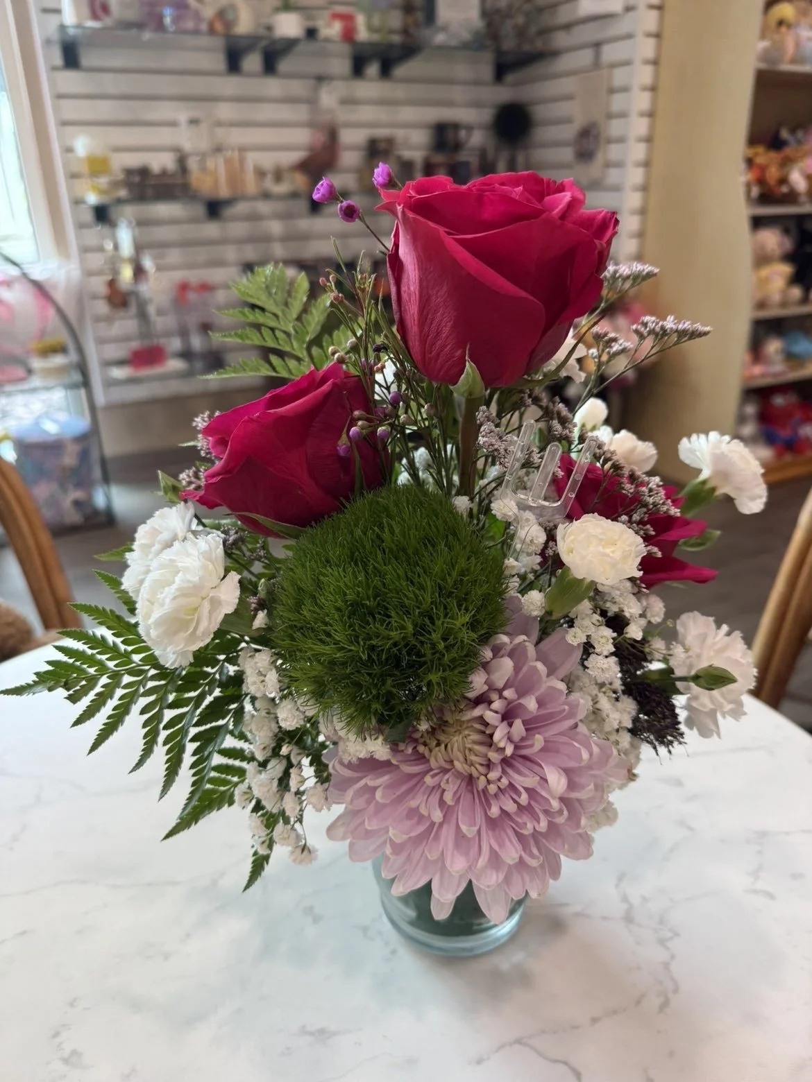 Un bouquet de fleurs avec des roses rouges, des chrysanthèmes roses, des œillets blancs, et d'autres petites fleurs, dans un vase en verre sur une table en marbre.